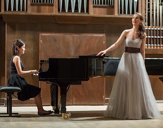 A woman stands before a piano and sings while another woman plays